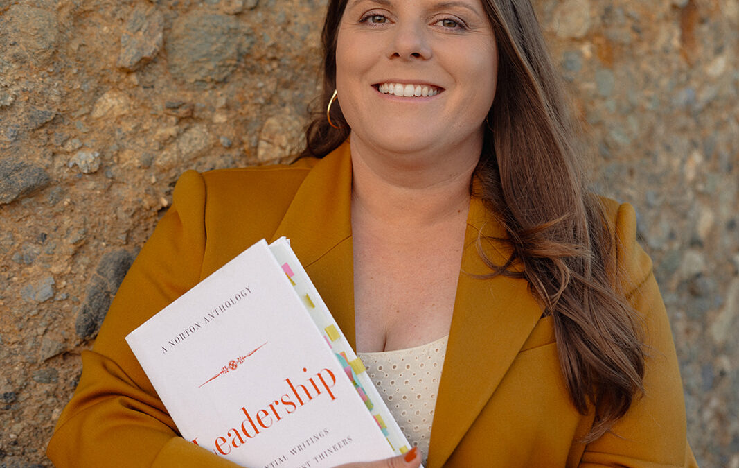 Liz Marion leans against a rock wall with desert tones, wearing a burnt orange blazer and holding a book titled "Leadership."