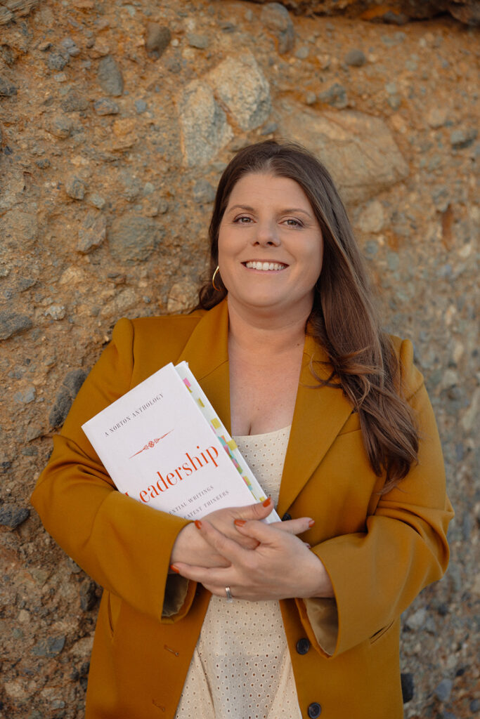 Liz Marion leans against a rock wall with desert tones, wearing a burnt orange blazer and holding a book titled "Leadership."